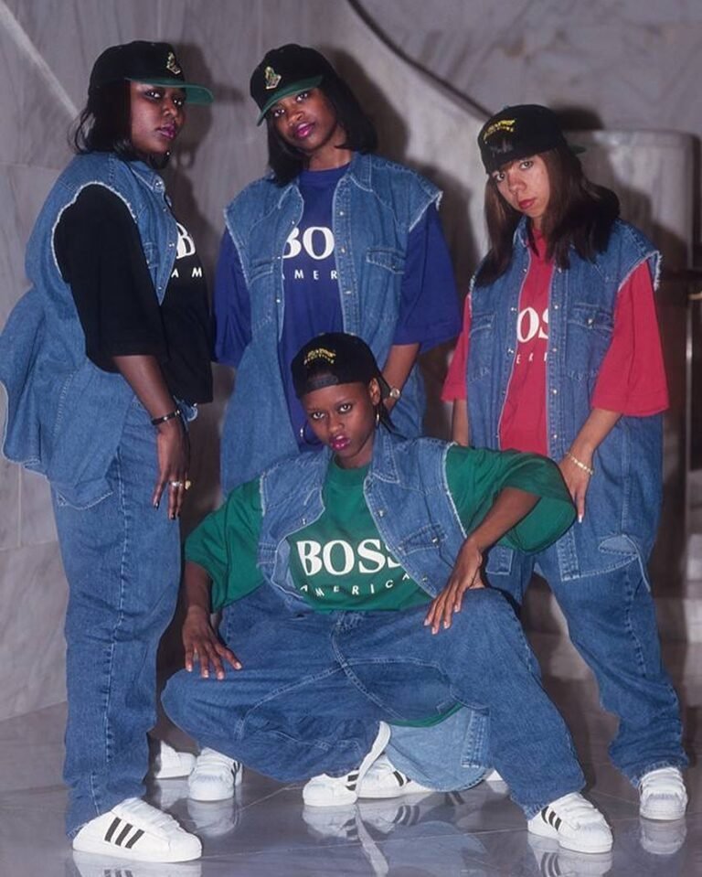 Grupo com camisetas largas, macacões jeans e bonés, referência do streetwear dos anos 1990.