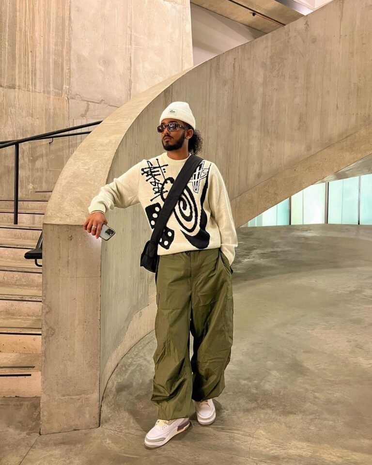 Camiseta larga estampada com calça verde e bolsa transversal em uma área urbana.