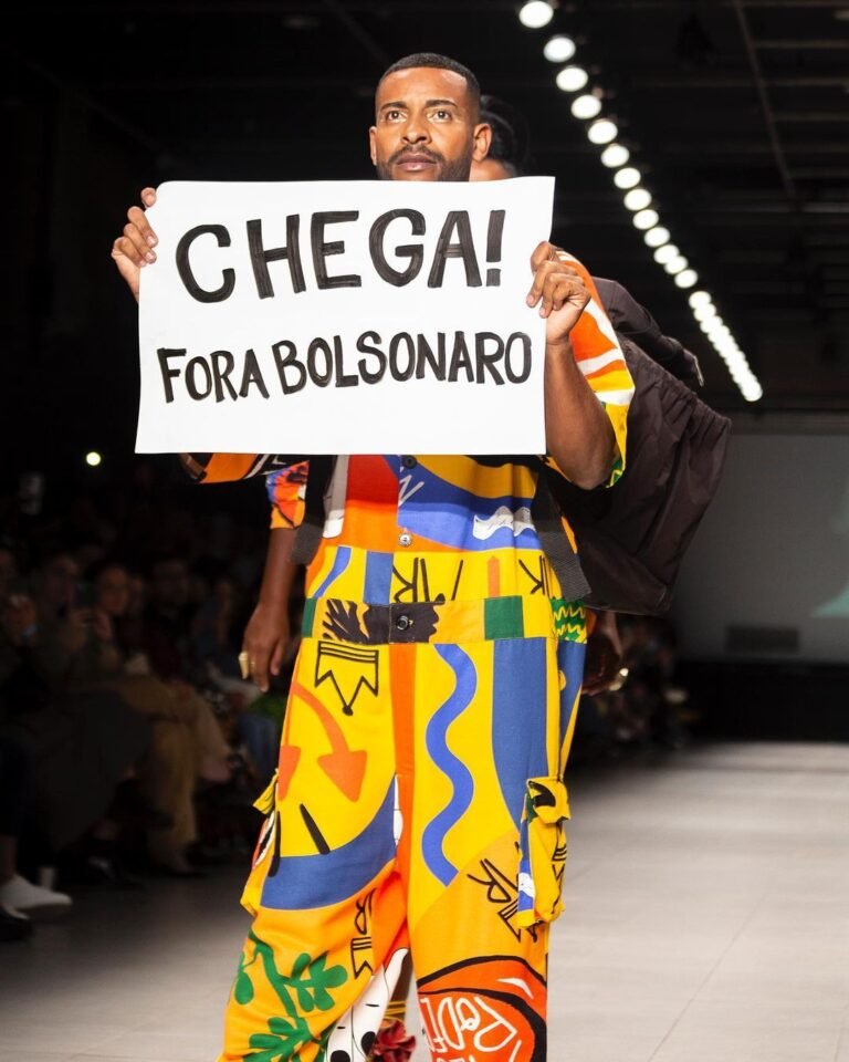 Modelo de roupa estampada segura o cartaz “Chega! Fora Bolsonaro” em uma passarela.
