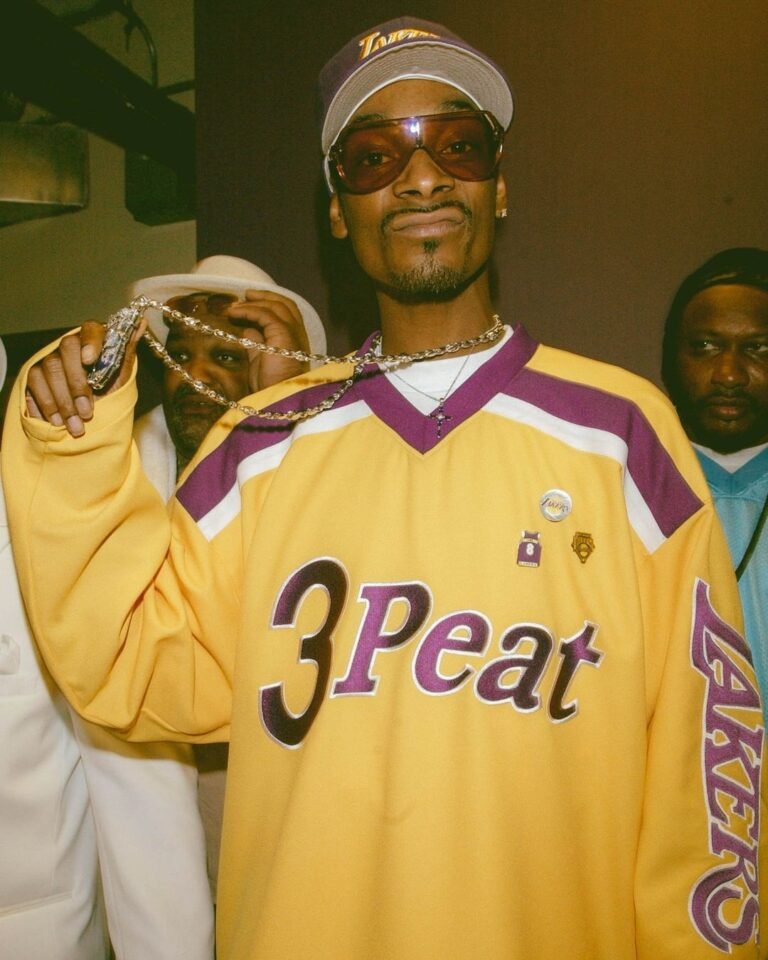 Snoop Dogg com camisa esportiva amarela, boné, óculos e colares.