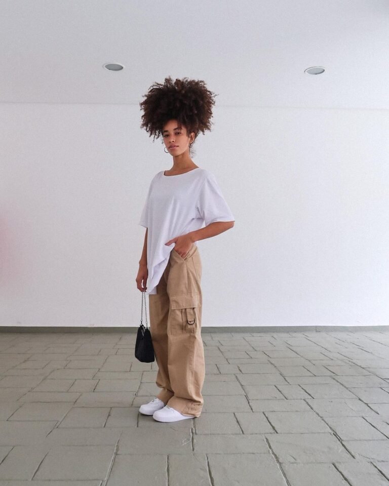 Camiseta branca ampla com calça marrom e tênis brancos em um ambiente claro.