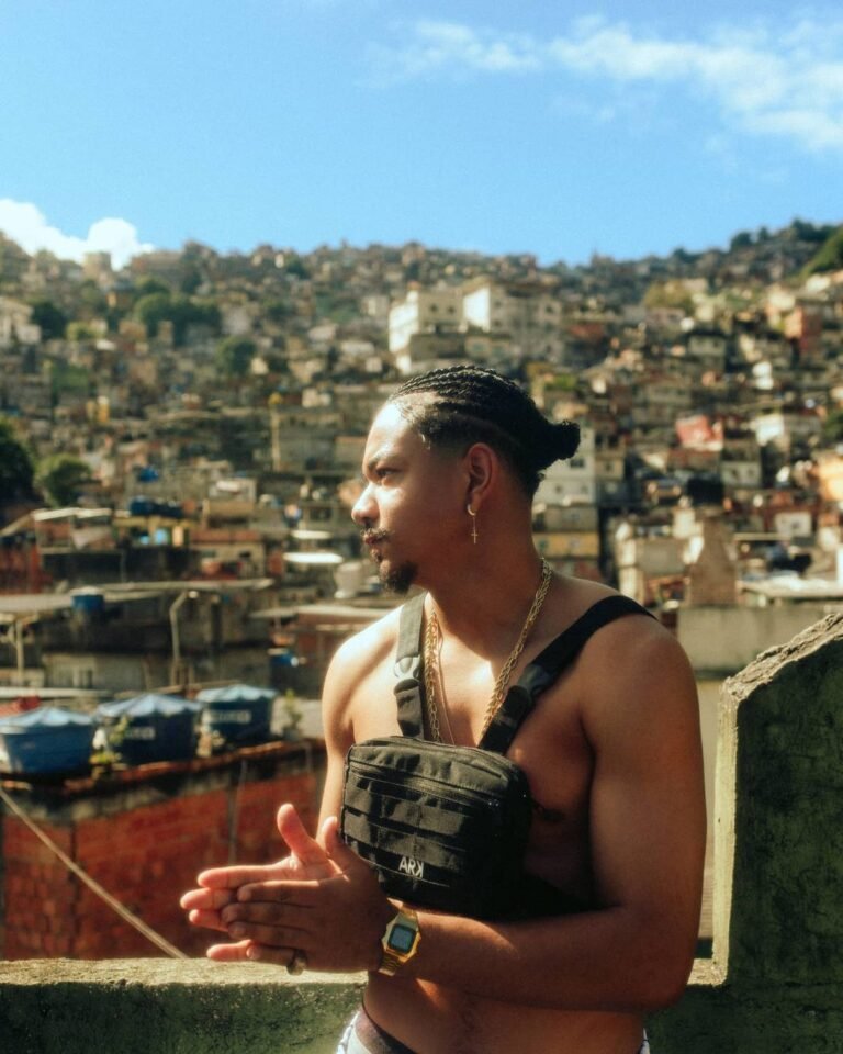 Cesar MC com bolsa transversal diante de uma vista de casas na encosta.