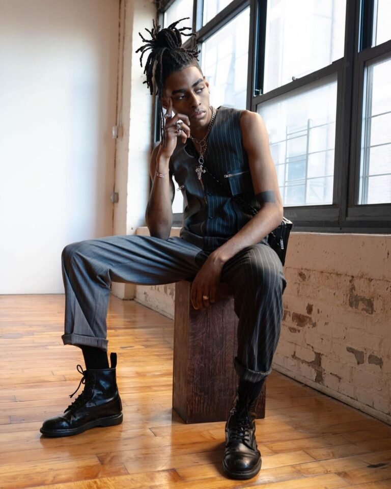 Look preto sem mangas com calça larga e botas, em um retrato sentado junto à janela.