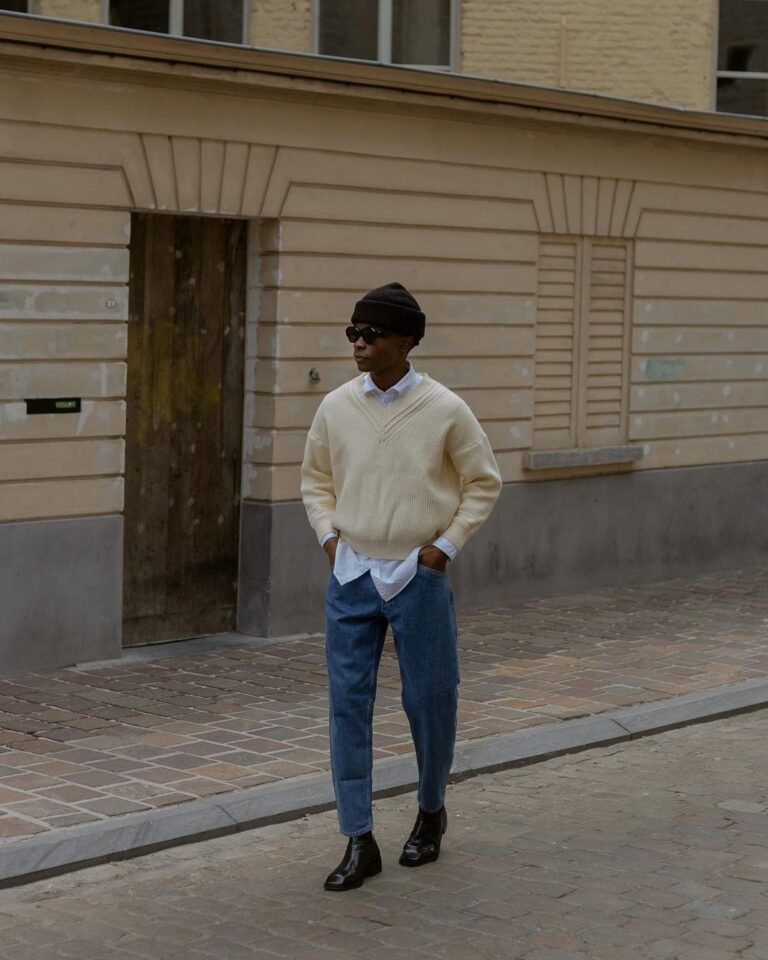 Suéter claro com calça jeans e botas pretas diante de uma garagem.