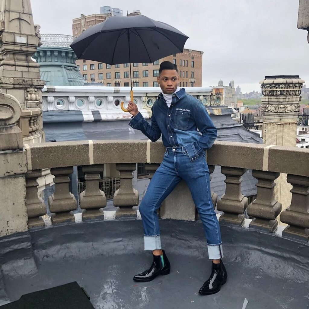 Homem com conjunto jeans azul, gravata e botas pretas segurando um guarda-chuva.