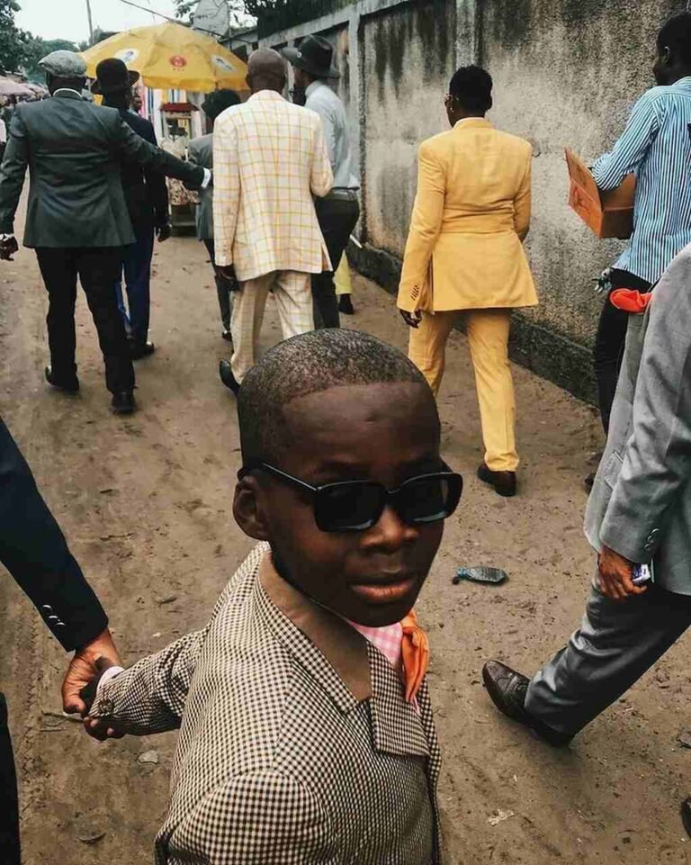 Criança de óculos e blazer em primeiro plano, com sapeurs ao fundo.