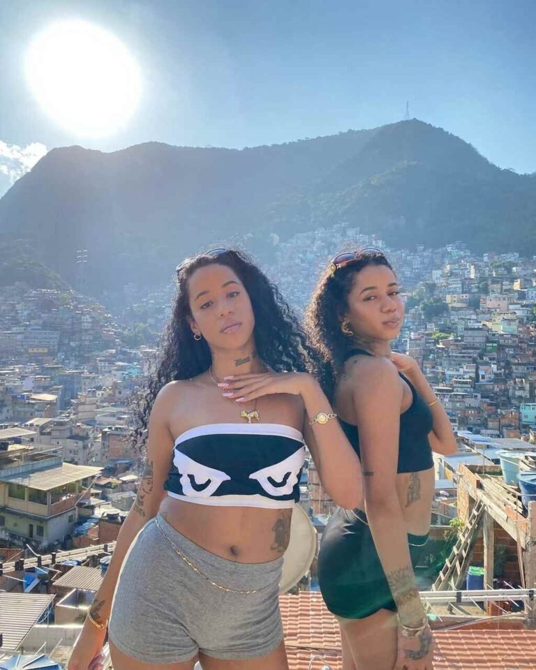 Tasha e Tracie com tops e shorts diante de uma vista de casas e montanhas.