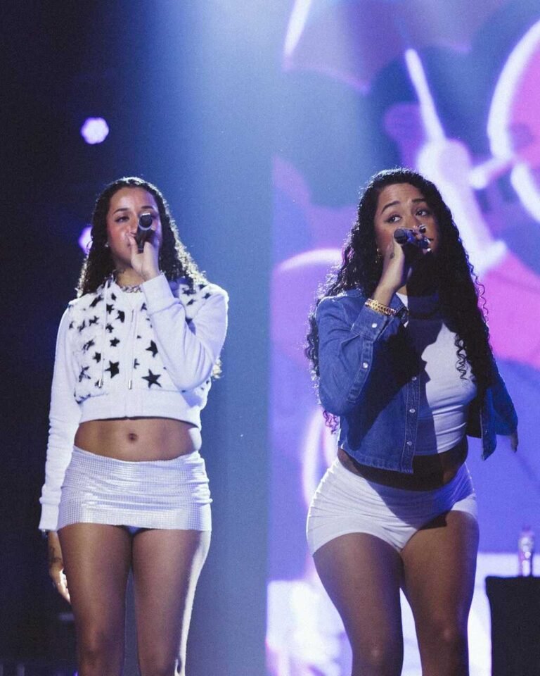 Tasha e Tracie no palco com top branco estampado, short e jaqueta azul.