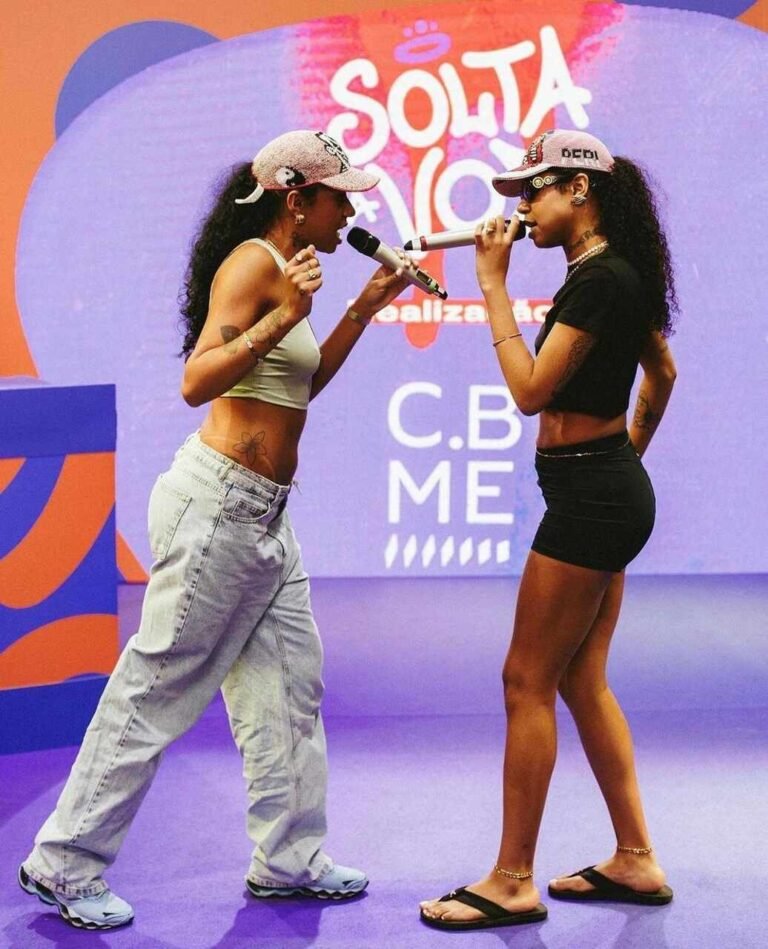 Tasha e Tracie cantam no palco de boné, top e calça larga ou saia curta.