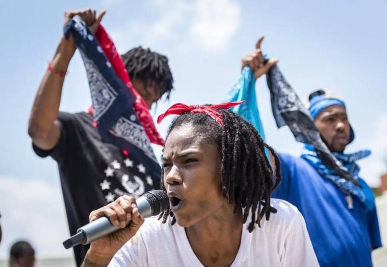 Pessoa com microfone durante manifestação, ao lado de bandanas azuis e vermelhas erguidas.